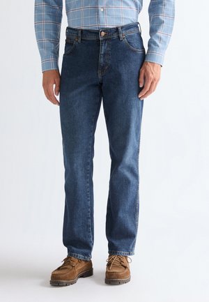 Mand iført blåternet skjorte, mørkeblå jeans med lige ben og brune snørestøvler, stående mod en ensfarvet hvid baggrund.