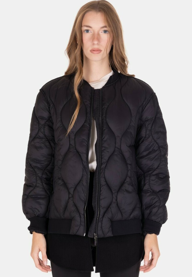 Giovane donna con lunghi capelli castano chiaro che indossa una giacca bomber trapuntata nera sopra una maglietta bianca, su uno sfondo semplice.