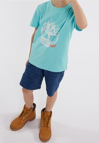 Niño con camiseta gráfica azul clara, pantalones cortos texturizados azul oscuro y grandes botas de cordones color beige de pie sobre un fondo blanco.