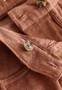 Ribikanga punases toonis riidest, millel on sooniline tekstuur, koos nuppude ja õmblustega. Lähedalt vaadates kattuvaid paneele ja metallkinnitusdetailide üksikasju.