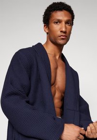 Navy waffle-textured bathrobe featuring a shawl collar, wide sleeves, and front pocket, showcased on a model with a bare chest.