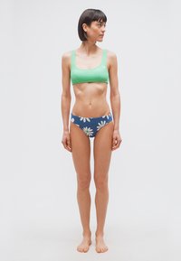 Γυναίκα όρθια ξυπόλητη, φοράει πράσινο αθλητικό σουτιέν και μπλε μαγιό με λευκά σχέδια μαργαρίτας, κοιτάζει προς τα αριστερά σε απλό φόντο.