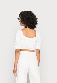 Blusa branca curta com mangas bufantes, design de costas quadradas e textura franzida. Combinada com calças de cintura alta de cor clara.