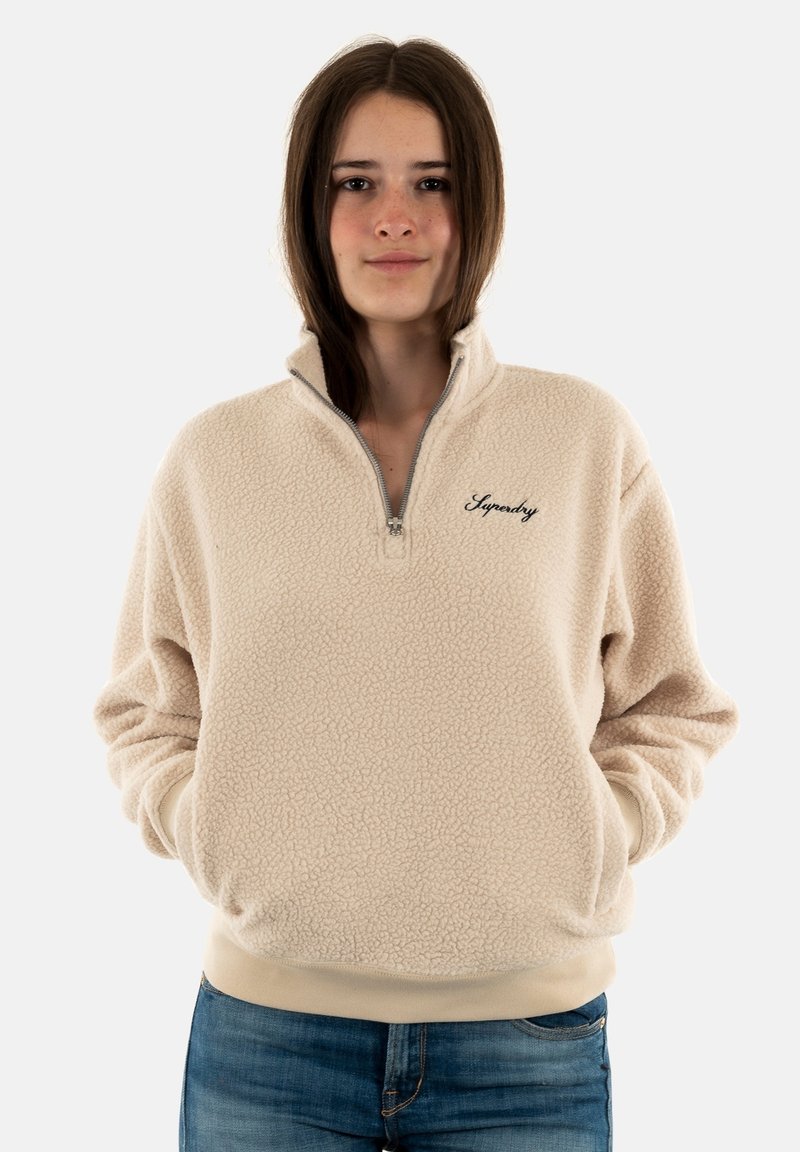 Sudadera de poliéster beige con media cremallera y puños acanalados. Presenta el nombre de la marca bordado en negro en el lado izquierdo del pecho. Textura acogedora.
