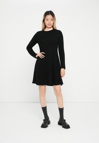 Vestido negro de manga larga con falda acampanada, con escote redondo y textura suave. Combinado con calcetas grises y zapatos negros.