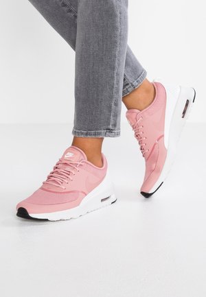 Voeten met roze Nike Air Max Thea sneakers met witte zolen en zwarte zooltips, gecombineerd met opgerolde grijze spijkerbroeken op een witte vloer.