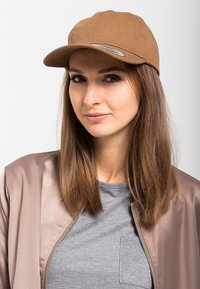 Bruine pet met een gebogen klep, gemaakt van stof. Heeft een zilveren accent op de klep en wordt gedragen met een grijs shirt en een beige bomberjack.