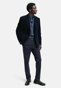 Marineblauer Cord-Blazer über einem Denim-Hemd, kombiniert mit dunklen Hosen und schwarzen Dressschuhen. Die glatte Textur und die taillierte Passform sind sichtbar.