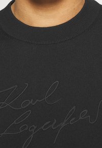 Schwarzes Sweatshirt mit geripptem Rundhalsausschnitt und strukturiertem Logodetail in derselben Farbe, das sich harmonisch mit dem Stoff verbindet.