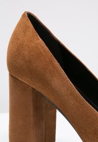 Talons hauts en daim marron avec un talon carré. L'empeigne présente une texture lisse, une pointe effilée et des coutures propres le long du bord.