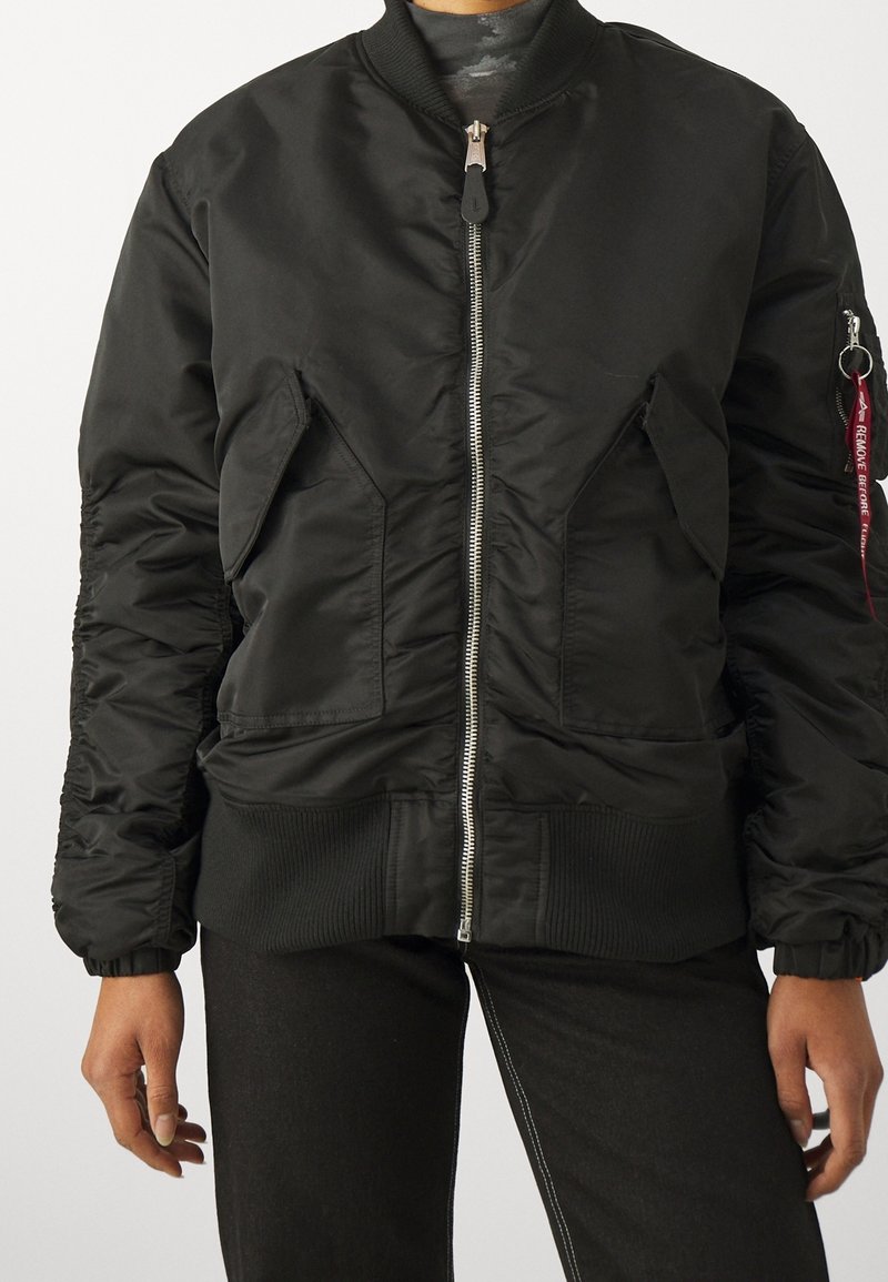 Personne portant une veste bomber noire avec une fermeture éclair sur le devant, des poches à rabat, des poignets côtelés et une étiquette rouge « Remove Before Flight » sur la manche.