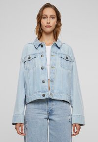 Chaqueta de mezclilla azul clara con un diseño recortado, cuello clásico, bolsillos frontales y cierres de botón. Presenta costuras visibles y un ajuste relajado.
