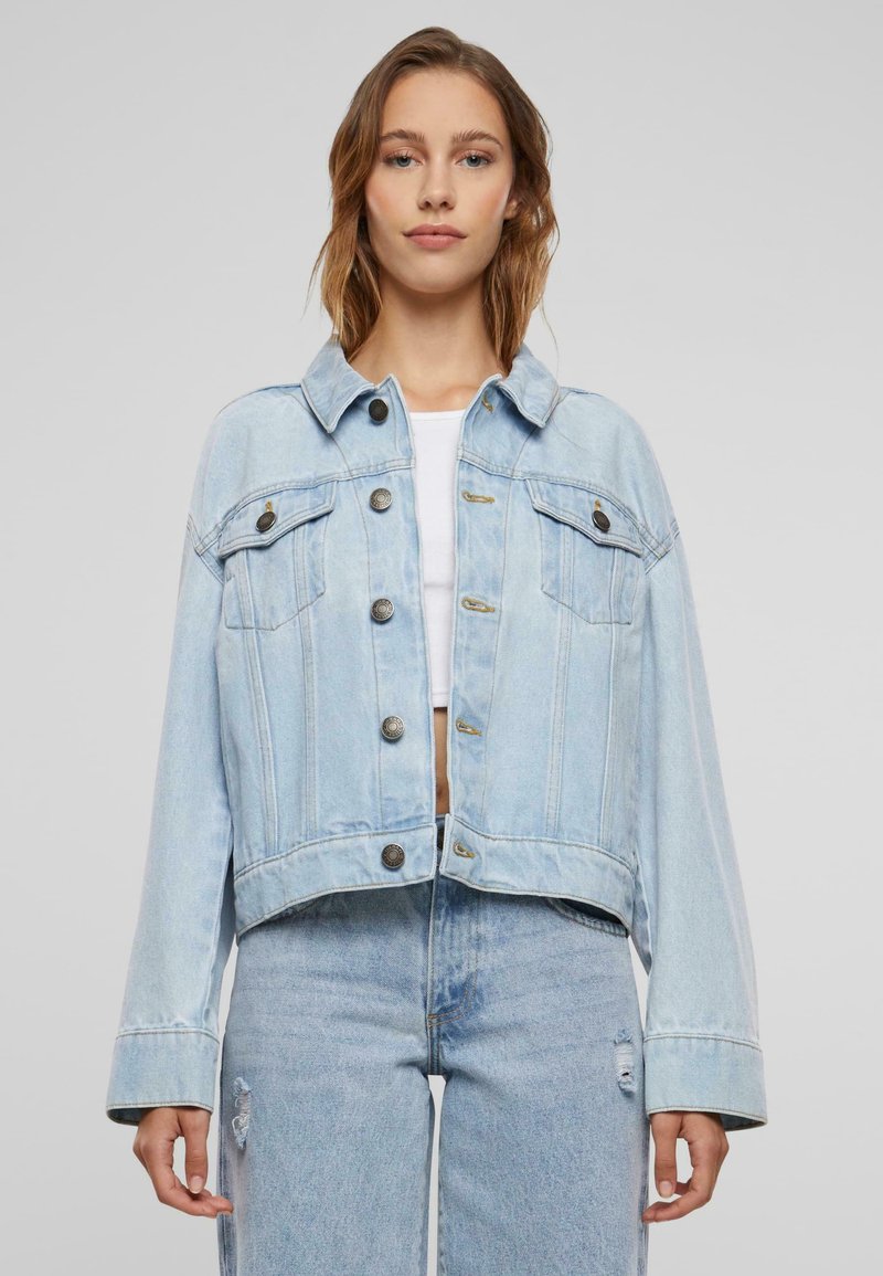 Chaqueta de mezclilla azul clara con un diseño recortado, cuello clásico, bolsillos frontales y cierres de botón. Presenta costuras visibles y un ajuste relajado.