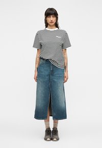 Μπλούζα με μαύρες και άσπρες ρίγες, δεμένη στη μέση, συνδυασμένη με μπλε τζιν μακρύ φόρεμα με σκίσιμο μπροστά και γκρι αθλητικά παπούτσια.