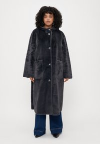 Manteau en fausse fourrure gris foncé avec une capuche, des boutons à l'avant et deux grandes poches latérales. Texture douce et design à coupe ample.