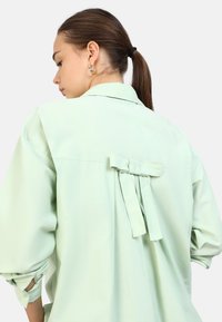 Camisa verde menta de tejido suave, con un detalle de lazo en la parte posterior y puños enrollados. Corte oversized con un diseño de cuello clásico.