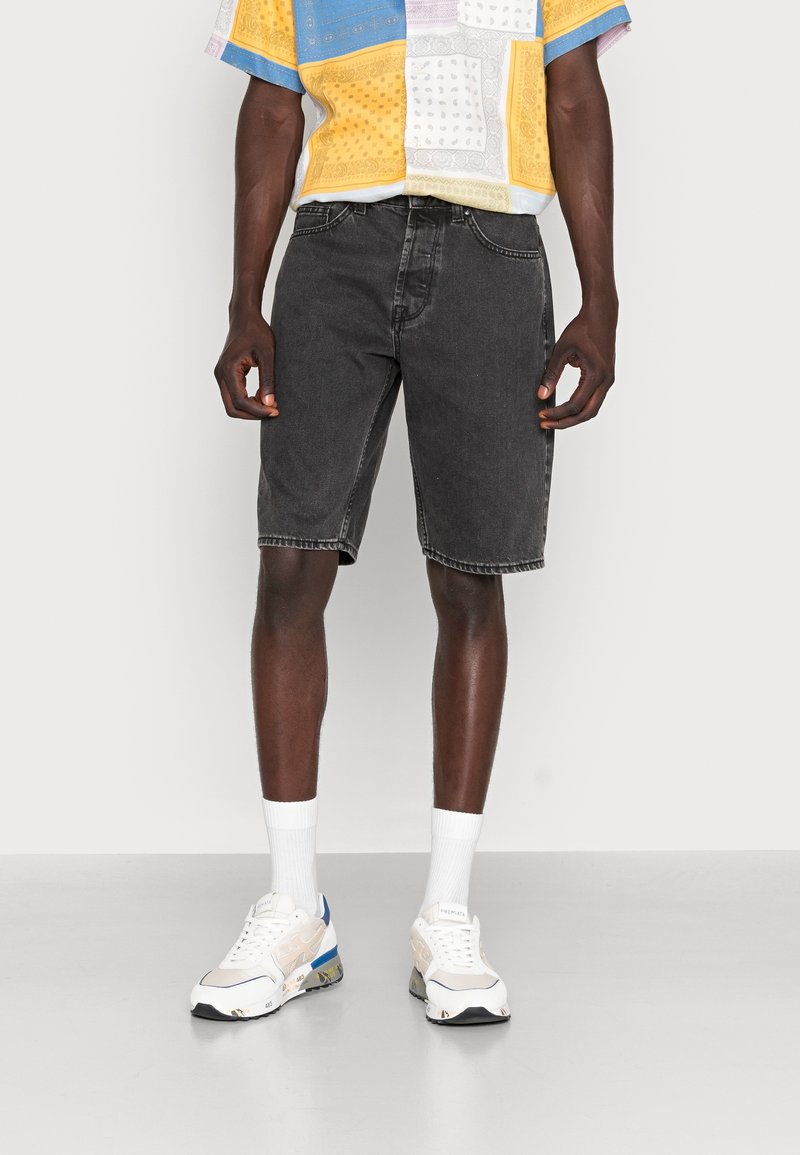 Bermudas de mezclilla negras para hombre, hasta la rodilla, con un ajuste relajado. Combinadas con una camisa de manga corta multicolor y de estampado, y zapatillas blancas.
