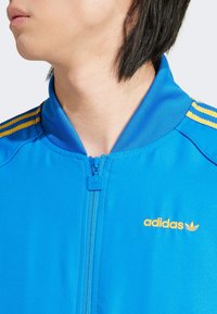 Plava jakna s patentnim zatvaračem izrađena od glatkog materijala, s tri žute pruge na ramenu i narančastim Adidas logotipom na prsima.