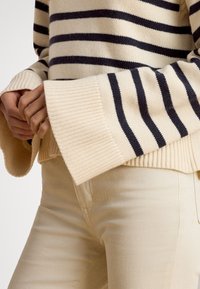 Kremfarget genser med marineblå horisontale striper, ribbestrikkede mansjetter og kant. Myk tekstur, kombinert med lyse beige bukser. Fokus på detaljene på ermet og mansjetten.