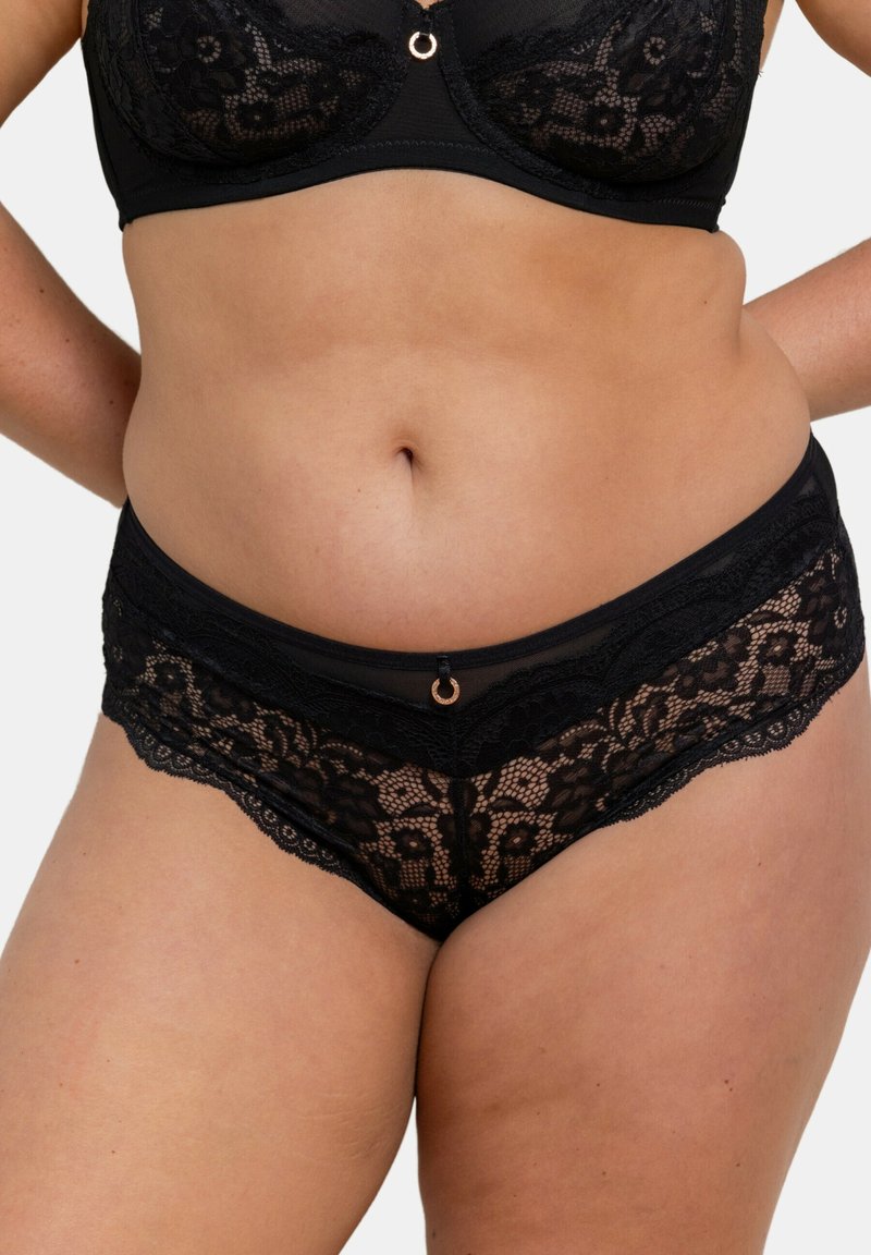Ensemble de lingerie en dentelle noire comprenant un bralette avec des bonnets transparents et un panty taille basse assorti. Détail en forme de cercle doré au centre devant.