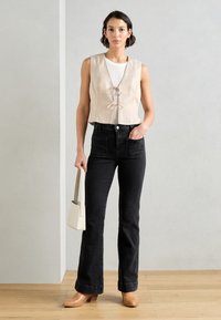 Beige linnen mouwloze top met een strikdetail aan de voorkant, gecombineerd met hoge zwarte flared jeans en tan kleurige hakken. Witte handtas.