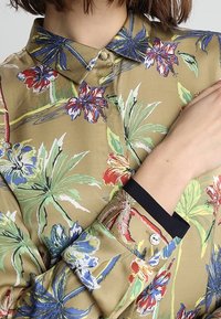 Blusa a fantasia floreale con sfondo khaki, maniche lunghe e polsini neri a contrasto. Design con colletto e chiusura a bottoni.