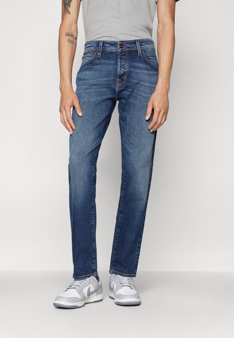 Jeans de mezclilla azul oscuro con un corte recto, que presentan un ligero desgaste y costuras en contraste, combinados con zapatillas gris claro.