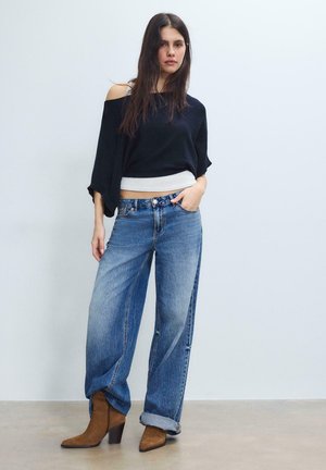 Donna con capelli lunghi e scuri che indossa un maglione nero scollato, una canottiera bianca, jeans a gamba larga blu e stivali con tacco marroni, in piedi all'interno.