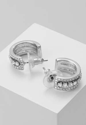 Boucles d'oreilles créoles en argent avec cristaux clairs incrustés et attaches en plastique transparent reposant sur une surface blanche.