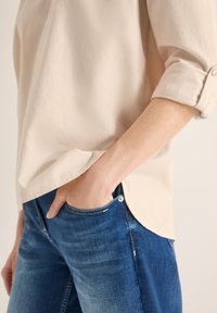 Vue de profil d'une personne portant une chemise beige à manches retroussées et un jean bleu, la main gauche reposant dans la poche avant du jean.