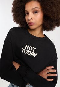 Felpa nera con colletto tondo e maniche lunghe, con la scritta "NON OGGI" in lettere bianche e in grassetto sulla parte anteriore. Tessuto morbido, vestibilità comoda.