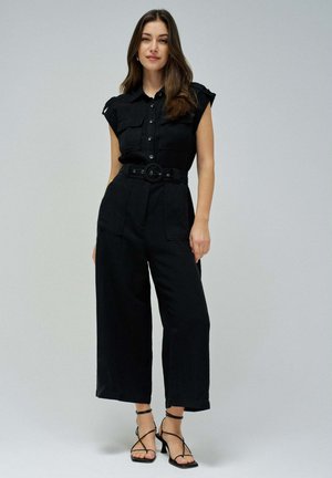 Schwarzer Jumpsuit aus Utility-Stil mit kurzen Ärmeln, Knopfleiste, tailliertem Gürtel und weitem Bein. Er hat zwei Vordertaschen und einen glatten Stoff.