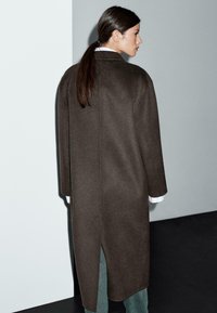 Manteau long en laine marron avec une coupe droite, des fentes sur les côtés et des épaules tombantes, superposé sur une chemise blanche et un pantalon vert.