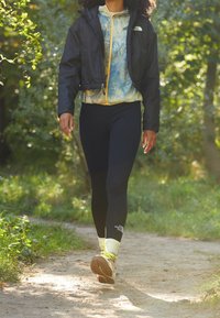 Beskuren svart vindjacka över en blå tie-dye zip-up, kombinerad med svarta leggings och ljusa vandringsskor med gula detaljer, promenerar på en stig.