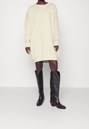 Persoon draagt een oversized beige sweatshirtjurk en zwarte leren kniehoge laarzen met roze accenten, staand op een lichte achtergrond.