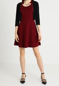Vestido burdeos tipo A con escote redondo, complementado por un cárdigan negro corto con ribete decorativo. Zapatos de tacón negro con tiras completan el conjunto.
