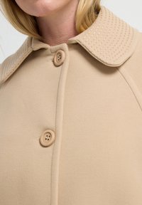 Beige jas met een gestructureerde, ribbelige kraag, voorzien van een knoopsluiting aan de voorkant en gladde stof. Twee ronde knopen zichtbaar aan de voorkant.