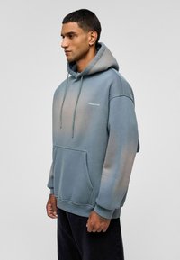 Hooded sweatshirt in een gradiënt van blauw-grijs, met een voorzak, trekkoord en een bedrukte logo "YOUNG ATELIER" op de borst. Zacht, gestructureerd katoen.