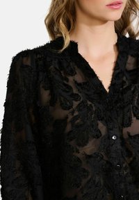Black, sheer blouse with textured floral embroidery, button front, and collar details. Soft fabric with a delicate, layered appearance.