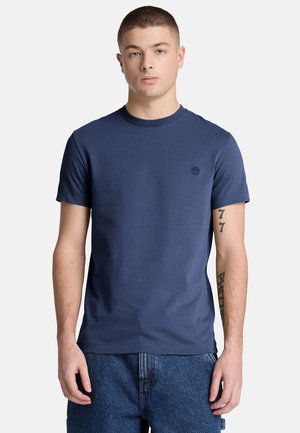 Uomo giovane che indossa una t-shirt semplice blu navy a maniche corte e jeans blu, in piedi davanti a uno sfondo chiaro con tatuaggi visibili sulle braccia.