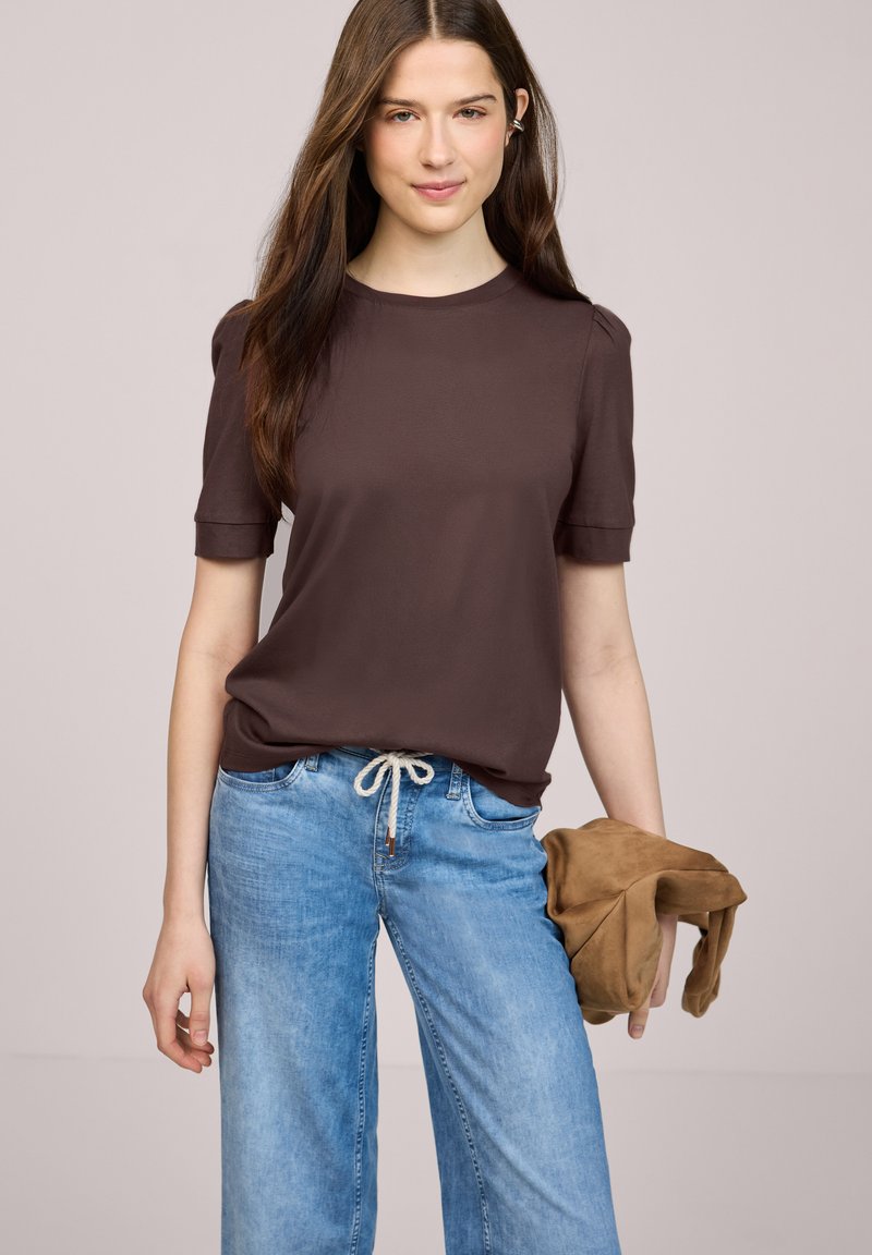 Ung kvinde med langt brunt hår iført en brun kortærmet skjorte og lyseblå jeans, der holder en sammenfoldet brun jakke i højre hånd.