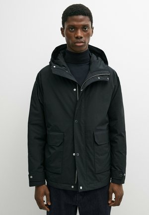 Junger Mann trägt eine schwarze Kapuzenjacke über einem schwarzen Rollkragenpullover und steht vor einem schlichten, hellen Hintergrund.