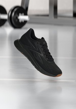Schwarzer Sportschuh mit brauner Sohle, der über einem Fitnessstudio-Boden schwebt, Fitnessgeräte im Hintergrund unscharf.