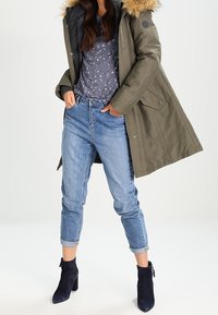 Parka verde oliva con capucha de piel, combinada con una camisa gris de flores y jeans de tiro alto en azul claro con los puños enrollados. Botas negras hasta el tobillo completan el conjunto.