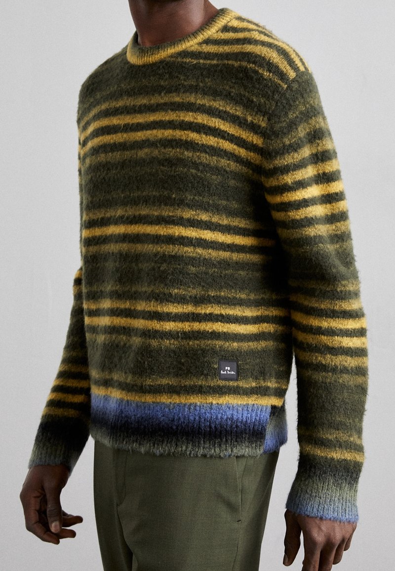 Homme portant un pull rayé duveteux aux tons jaune, vert et bleu, associé à un pantalon vert olive, vu du cou aux hanches.