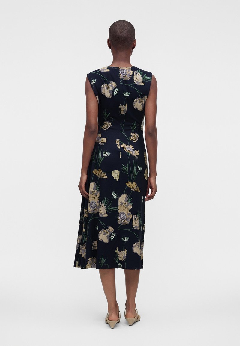Person standing with back to camera wearing a sleeveless, dark floral dress and beige heels against a plain, light background.