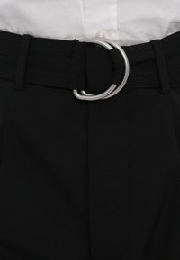 Pantaloni din țesătură neagră, cu un talie lată și o cataramă circulară argintie. Textura este netedă, cu detalii subtile de pliere.