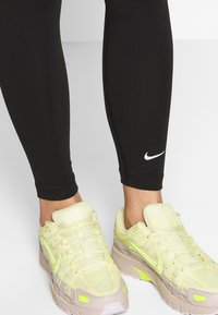 Svarta leggings med en vit Nike-logga nära vristen, kombinerat med ljusgula Nike-sneakers med grå detaljer och snören.