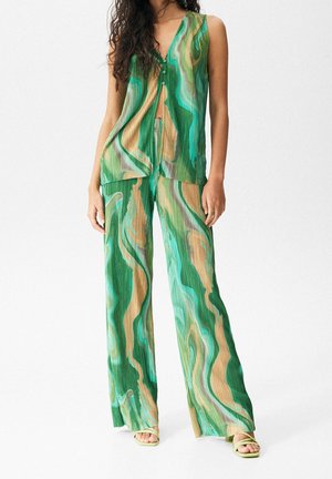 Femme portant un haut sans manches boutonné aux motifs ondulés verts, beiges et bleus, et un pantalon large assorti, avec des sandales vert clair.