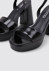 Tacones de plataforma negros con un acabado brillante, tira frontal ancha y tacón alto cuadrado. Diseñados con la punta abierta y correas delgadas en el tobillo.